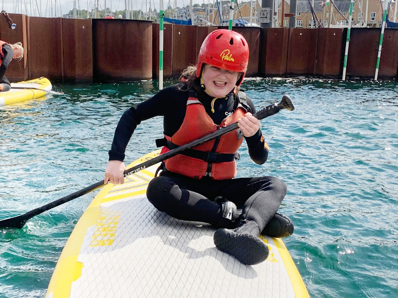 Katie wearing a black wetsuit, and red life jacket and hard hat. She's sat on top of a paddle board and holding a paddle. She is grinning broadly and looks like she's having a brilliant time!