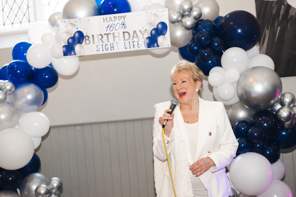 Picture of Beverley Humpreys singing in front of balloon arch with Happy 160th Birthday Sight Life sign at top.