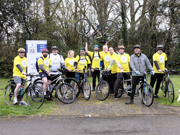 Picture of eleven cyclists sat on their bikes with one in the centre holding the bike above his head.